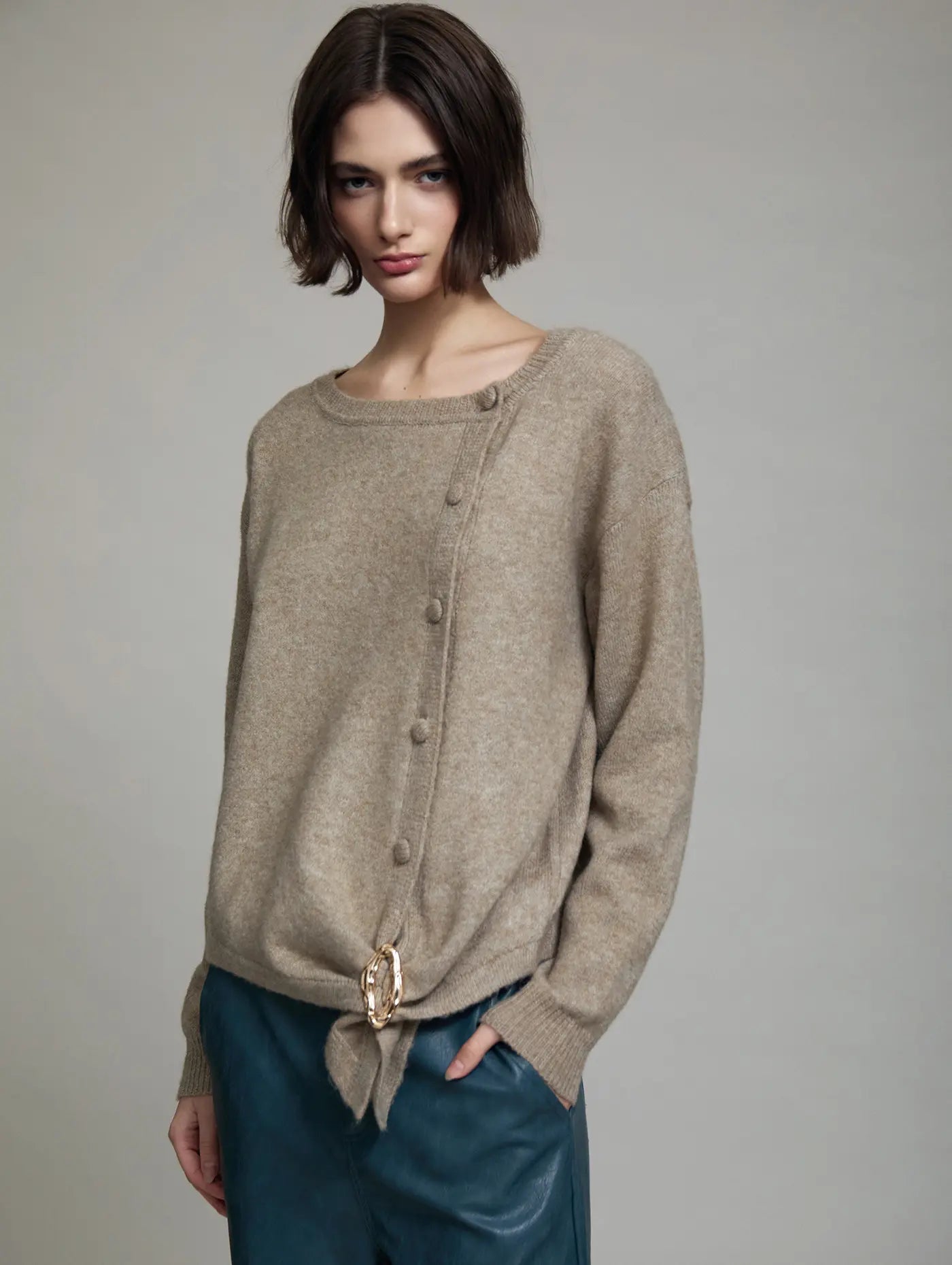 Cardigan Sienna de SKATIE en punto beige mélange con manga larga, botonadura lateral en diagonal, bajo asimétrico efecto pañuelo y broche metálico dorado