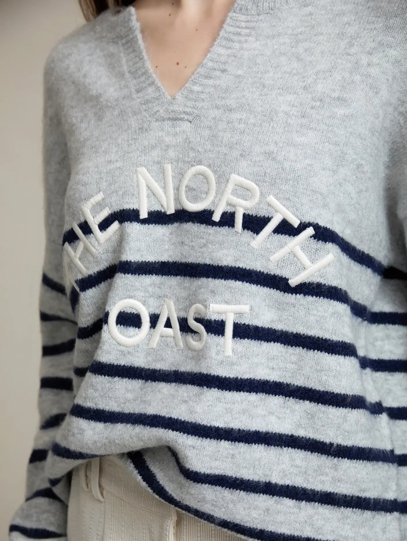 Detalle del Jersey The North Coast de SKATIE en gris claro jaspeado con rayas azul marino, escote en V y bordado frontal en relieve The North Coast en color crudo